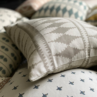 A close-up of a cushion, lying amongst a pile of other cushions that are out of focus. The main cushion has a bold pattern of diamonds in natural and cream. It sits on another cream cushion with a small floral repeat pattern.