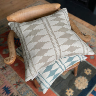 A photo of two cushions from above, sitting on a wooden armchair with a patterned green and rusty red rug beneath. The cushions both have a old diamond shape pattern, the top one in natural colours and the bottom one in green blue and cream.