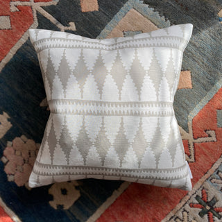 A picture from above of a square cushion lying on a dark teal and rusty red patterned rug. The cushion is in dappled sunlight and has a bold diamond shaped pattern in light natural and cream colours.