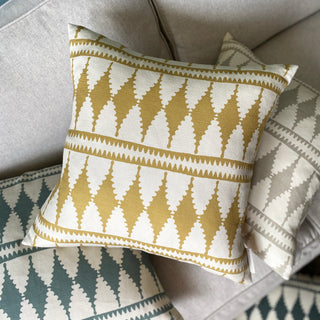 A picture of a cushion from above, lying on a pile of cushions on a pale grey sofa. The cushions are all in the same bold diamond pattern, the main cushion being ochre and cream. The cushions below are slightly out of focus, in a slate green and natural.