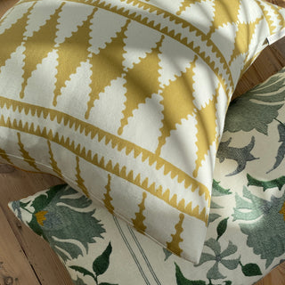 A close up of a the corner of a cushion in dappled sunlight, lying on a wooden floor on top of another cushion. The main cushion has a bold diamond shaped pattern in ochre and cream, with the cushion underneath having a striking floral suzani pattern in blue green.