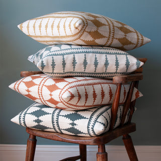 A pile of four cushions on a wooden armchair, infront of a deep blue coloured wall. All of the cushions have a bold diamond pattern with cream background. The colourways from the bottom up are slate green, terracotta red, green blue and natural at the top.