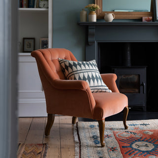 A pinky red velvet armchair sits in front of a fireplace with a wood burning stove and a wooden mirror over the mantlepiece.  The armchair has a bold diamond patterned cushion on it, in slate green and cream. There's a patterned rug underneath the armchair in terracotta red and teal blue.