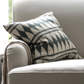 A square cushion leans on the armrest of an oatmeal armchair with a throw hanging over the armrest. The cushion has a bold cream and natural diamond pattern. The armchair is sitting in front of a window.