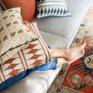 A close-up image of someone sitting on a grey linen sofa, only their legs and feet in shot. They are holding two patterned cushions on their lap, the bottom one in a bold terracotta and cream diamond pattern and the top in a floral stripe motif pattern in cream, pink and green. There is a terracotta and teal patterned rug on the floor.