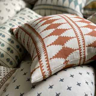 A close-up of a square cushion, lying amongst a pile of other cushions that are out of focus. The main cushion has a bold pattern of diamonds in terracotta and cream. It sits on another cream cushion with a small floral repeat pattern.