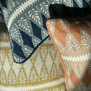 A close-up of the corners of three cushions from above, all positioned at different angles. They all have the same detailed pattern with a zig zag stripe and scalloped pattern. The top one is pink and cream, the middle one is dark blue, grey and cream and the bottom one is ochre and cream. Sunlight is falling to the left of the pile, creating shadows to the right.