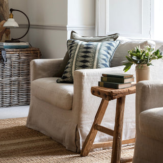 A linen armchair sits in front of a wicker basket  with a lamp on top and next to a wooden stool with books and flowers on it. The armchair is skirted with two cushions leant against the armrest, the front one with a detailed pattern in blue and cream. There is light coming in from the window behind and the armchair sits on a natural jute rug.