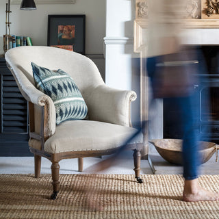 A curved back natural linen armchair with wooden legs sits next to a fireplace with wood burning stove and infront of a black cupboard. The armchair has a patterned blue and cream cushion sitting on it, and there is a blurred figure walking across the  room in the foreground of the picture. There is a natural jute rug underneath the armchair.