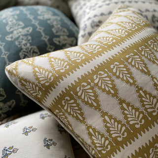 A close-up of the corner of a patterned cushion in ochre and cream. The cushion has a zig zag stripe and scalloped pattern. There are other cushions out of focus in a pile underneath it.