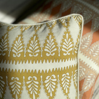 A close up of the corner of a cushion with sunlight shine on it. The cushion has a detailed zig zag pattern and is in an ochre and cream colourway.  There is another cushion out of focus in the background in a pink colour.