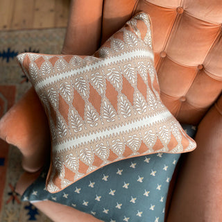 A shot from above of a square patterned cushion in rusty pink, nude and cream. The pattern is detailed, with a zig zag stripe and scalloped floral repeat. The cushion lies on top of a slate green cushion with a small ditsy repeat, both of which are lying on terracotta velvet armchair.