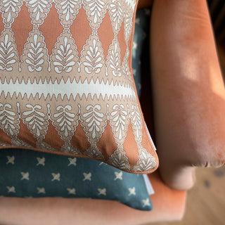 A close up of the corner of a square patterned cushion in rusty pink, nude and cream. The pattern is detailed, with a zig zag stripe and scalloped floral repeat. The cushion lies on top of a slate green cushion with a small ditsy repeat, both of which are lying on terracotta velvet armchair.