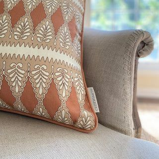 A close up of the corner of a square patterned cushion in rusty pink, nude and cream. The pattern is detailed, with a zig zag stripe and scalloped floral repeat. The branded Clothsmith label is visible in the bottom corner. The cushion sits on a natural linen armchair, with exposed stitching on the arm.