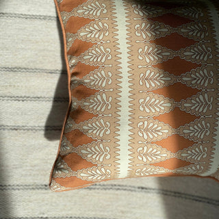 A shot from above of the corner of a square patterned cushion in rusty pink, nude and cream. The pattern is detailed, with a zig zag stripe and scalloped floral repeat. The cushion lies on a cream striped rug, with sunlight falling across half of it.