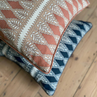 A close up shot from above of a square patterned cushion in rusty pink, nude and cream. The pattern is detailed, with a zig zag stripe and scalloped floral repeat. The cushion lies on top of a dark blue cushion in the same pattern which is out of focus, both of which are lying on a stripped pine wooden floor.