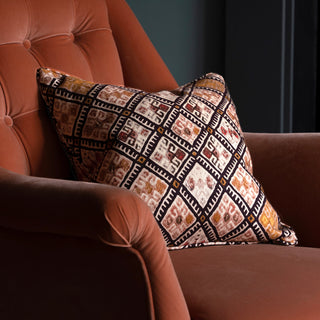 A geometric patterned cushion in black, pink and ochre, sits on a terracotta velvet armchair with buttons. Behind the armchair is a dark green wall.