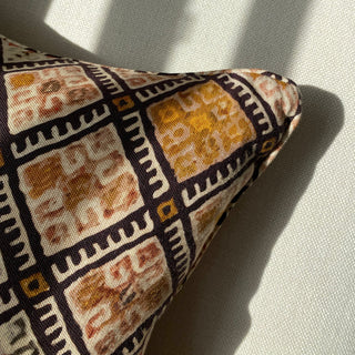 A close up of the corner of a cushion with a geometric pattern in black, pink and ochre. It lies on a cream textured armchair, with sunlight falling across it creating shadows.