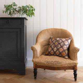 A geometric patterned cushion in black, pink and ochre, sits on an antique French armchair with with turned wooden legs. Behind the armchair is a cream panelled wall and next to it is a black wooden cupboard with green foliage sitting on top of it.
