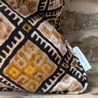 
A close up of the corner of a cushion with a geometric pattern in black, pink and ochre. The branded Clothsmith label can be seen in the bottom corner. It sits against an exposed stone wall.