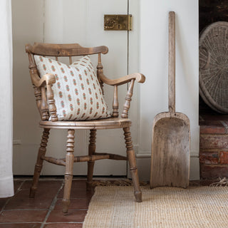 A cream striped square cushion with a pink floral motif sits on a wooden armchair in the corner of a dining room, with a tiled teracotta floor and jute rug underneath. An antique wooden shovel is leaning up against the wall next to the chair.