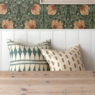 Two cushions sit on a bench leaning against cream wooden panelling with a floral green and peach wallpaper above. The cushions are patterened, one in green blue and cream, the other with a pink foral stripe on cream background. There is a wooden table in the foreground.