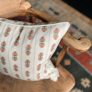 A shot from above of a square patterned cushion with a pink floral stripe on a cream background. The cushion sits on a wooden armchair with a patterned green and red rug beneath it.