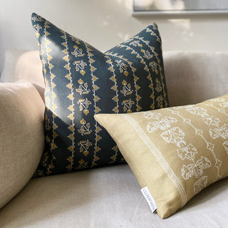 Two cushions sit on a natural linen sofa with dappled sunlight falling across them. The larger square one is in a dark slate green with an detailed ochre stripe pattern. The smaller rectangular cushion leans up against it and is in a soft hay yellow with a branded Clothsmith label.