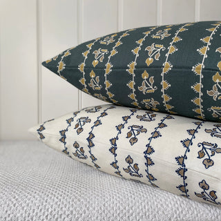 A close up of the corner of two cushions, one on top of the other, sitting on a textured cream throw wiith a cream panelled wall behind. The cushions have the same detailed stripe, the top one being a dark slate green and the bottom one is cream.