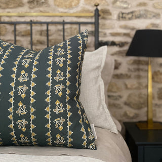 A square cushion sits to the left of the image on a bed with white bed linen. The bed is out of focus and sits in front of an exposed stone wall. The cushions has a detailed stripe in ochre on a slate green background. A branded Clothsmith label can be seen in the corner.