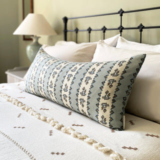 A cream-colored lumbar cushion with a geometric indigo blue and gold stripe pattern, placed on a bed next to a black headboard and a lamp.