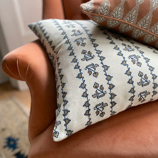 A square cream cushion with detailed pattern in a blue stripe lies on a terracotta velvet armchair with the corner of a pink patterned cushion in the top corner.