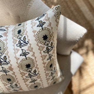 A square cushion cover with a floral pattern in soft pink and natural tones, featuring a heritage style design sitting on a natural linen armchair in sunlight.
