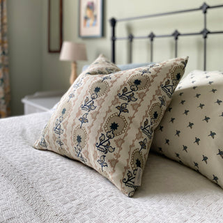 A square cushion cover with a floral pattern in soft pink and natural tones, featuring a heritage style design on a bed with cream bedspread.