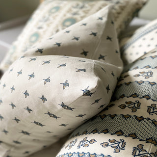 A close up of a square cushion with a natural background featuring a small scale repeat of green and gold flower motifs.