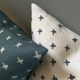 A close up of two square cushions with featuring a small scale repeat of green and gold flower motifs. One is cream the other is dark slate green.