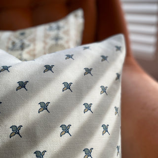 A close up of a square cushion with a natural background featuring a small scale repeat of green and gold flower motifs, with light falling across the cushion.