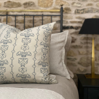 A square cushion with an intricate dark blue floral line drawing on a natural linen background is placed on a bed with a stone wall and lamp in the background.