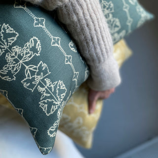 A slate green square cushion cover with an intricate off white line drawing design, held underneath someone's arm with a yellow cushion behind it.