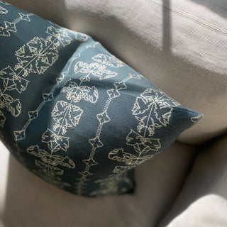 A slate green square cushion cover with an intricate off white line drawing design, sitting on a natural linen sofa in the sunshine.