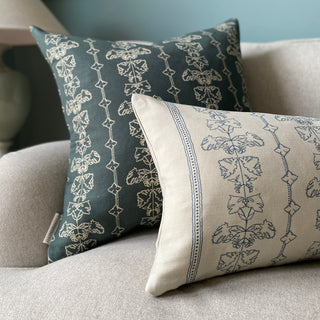 A slate green square cushion cover with an intricate off white line drawing design, sits behind a natural linen rectangular cushion in the same design.