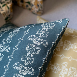 A slate green square cushion cover with an intricate off white line drawing design, lies on a yellow cushion in the same design.